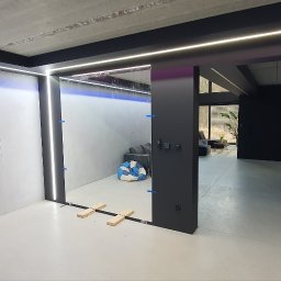 DKHOUSE - Nowoczesne wnętrze w trakcie wykończenia: surowy beton, minimalistyczne oświetlenie LED, czarne elementy konstrukcyjne i widoczna osoba w tle.