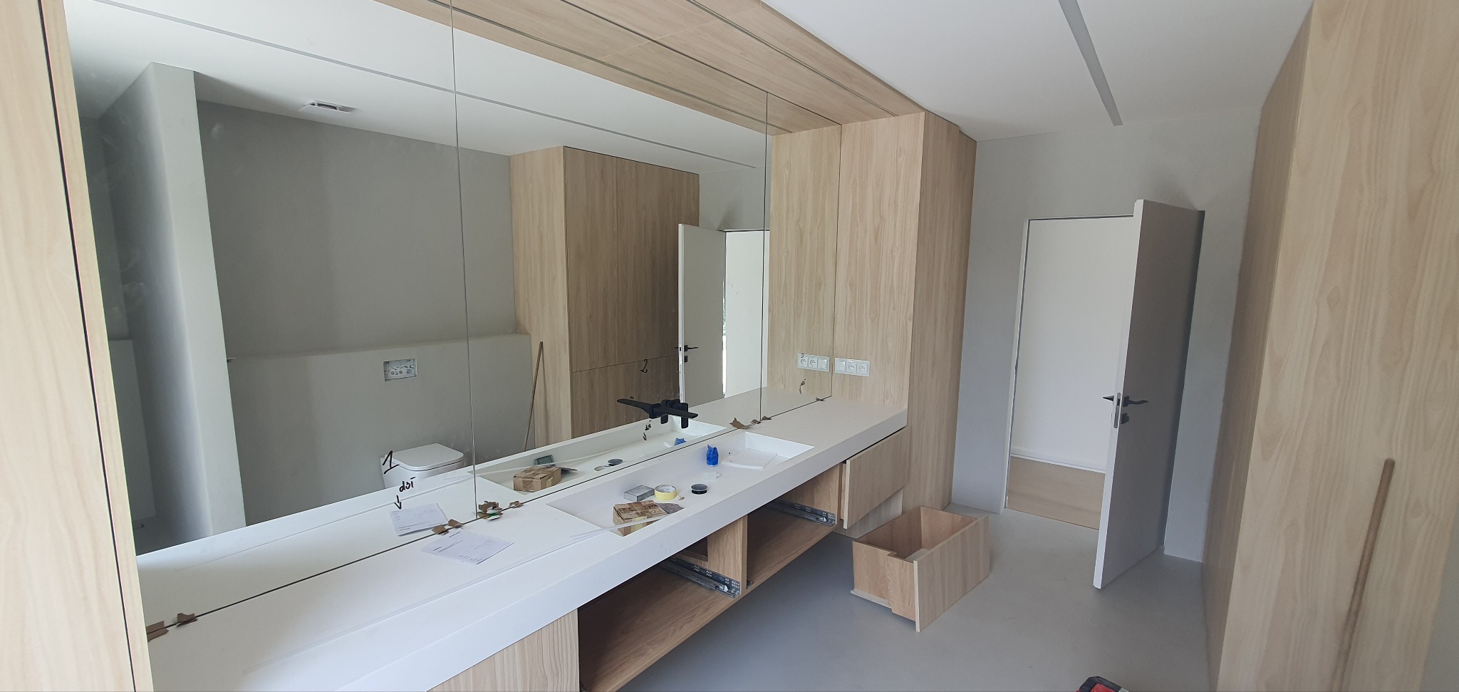 Nowoczesna łazienka w trakcie montażu: biała umywalka z szafką z jasnego drewna, duże lustro z widocznym odbiciem toalety. Elementy wykończeniowe w trakcie instalacji.
