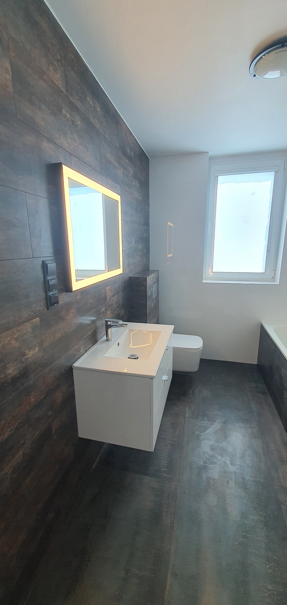 Nowoczesna łazienka z ciemnymi płytkami imitującymi drewno, białymi sanitariatami i podświetlanym lustrem. Minimalistyczny design, jasne światło z okna.