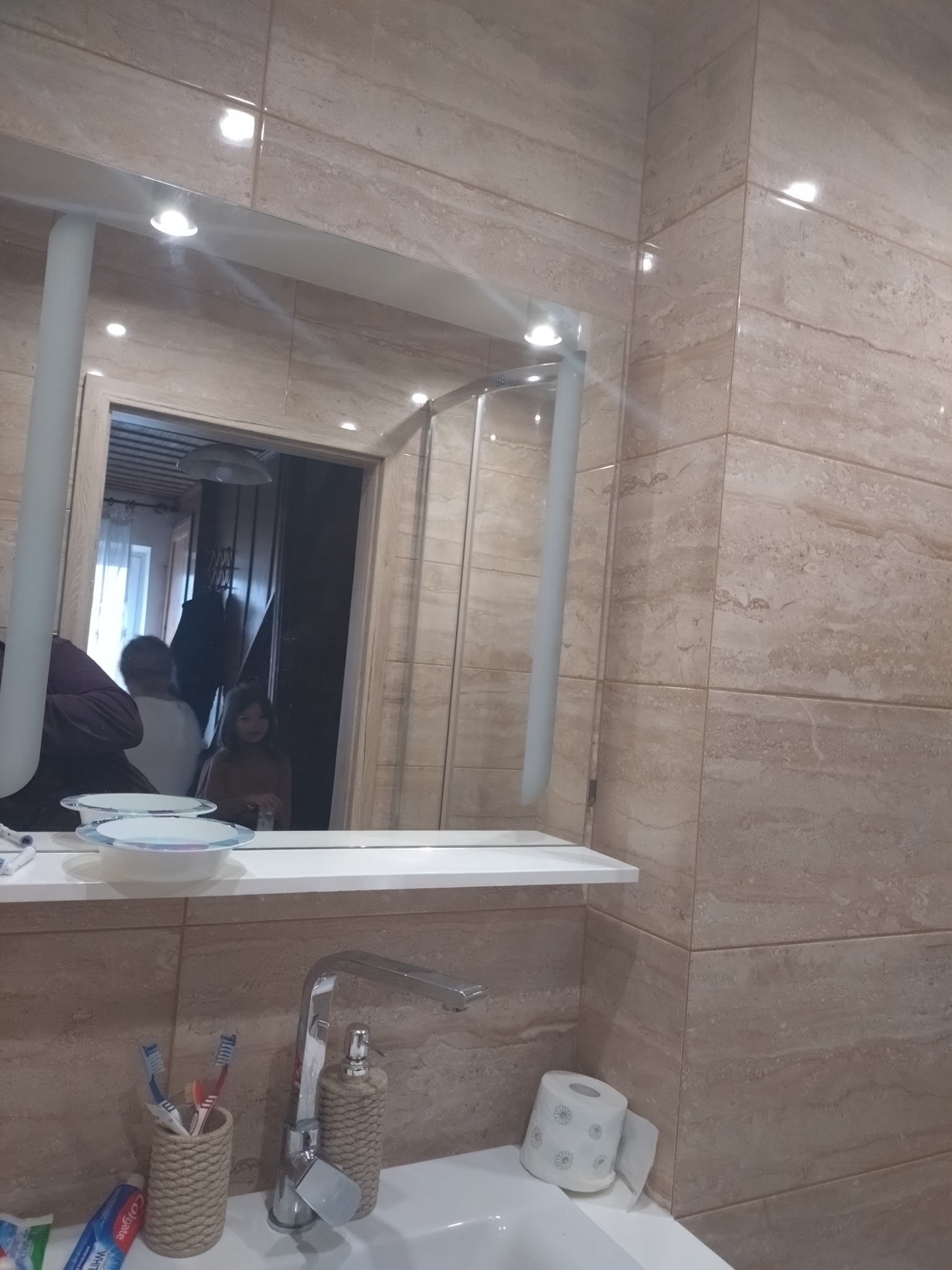 Oświetlenie punktowe w łazience z beżowymi płytkami, odbijające się w lustrze nad umywalką. Widoczne akcesoria łazienkowe i fragment wnętrza w odbiciu.