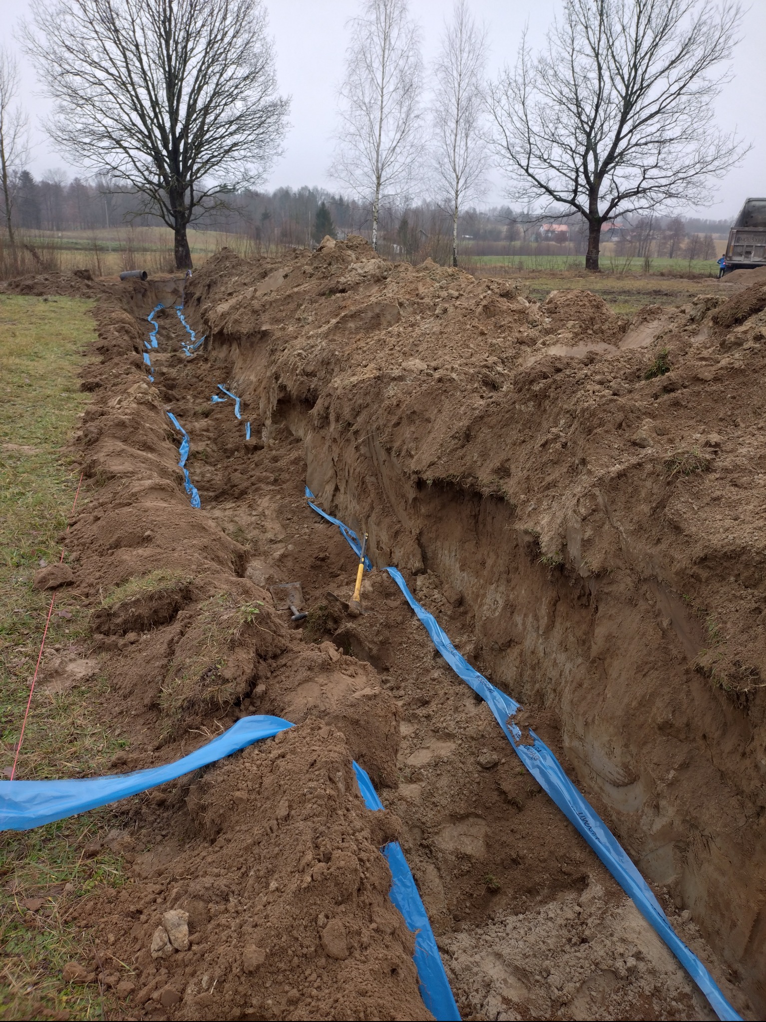 Wykop z ułożonymi niebieskimi rurami w ziemi, prawdopodobnie pod instalację elektryczną. Widoczne narzędzia i ziemia. Tło stanowią drzewa i pochmurne niebo.