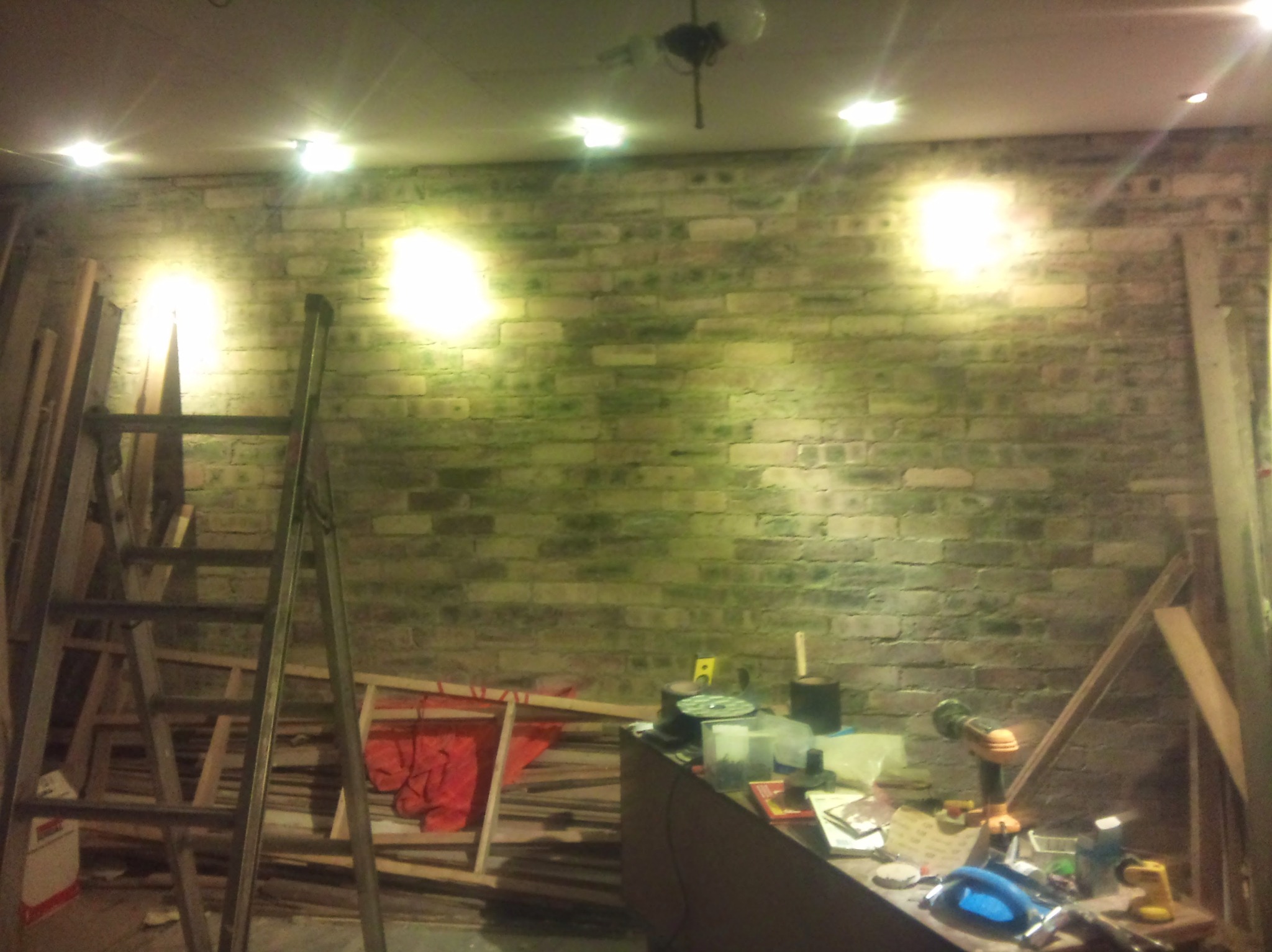 Ściana z cegły oświetlona punktowymi lampami sufitowymi. Widoczna drabina, narzędzia i materiały budowlane. Etap prac remontowych, surowy stan wykończenia.