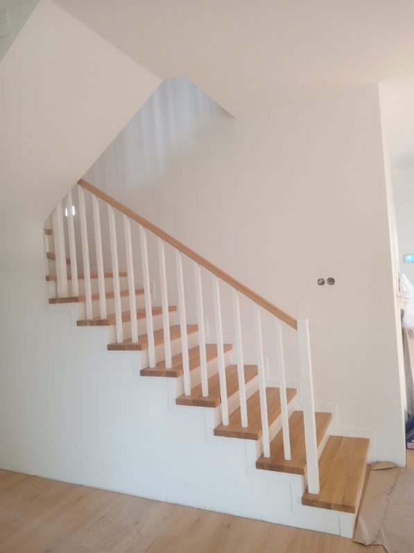 Jasne schody z drewnianymi stopniami i białą balustradą w minimalistycznym wnętrzu. Widoczne gniazdka elektryczne na ścianie.