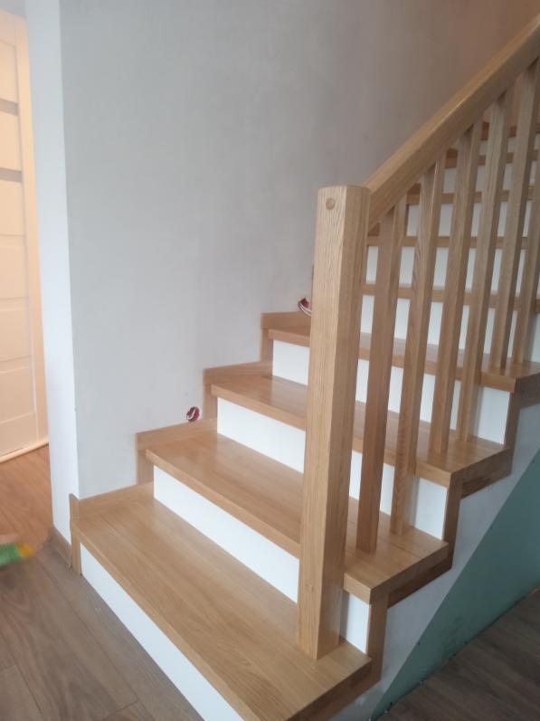Jasne, dębowe schody z białą podbitką i prostą balustradą. Widok z dołu, ukazujący detale wykończenia i naturalne usłojenie drewna. Minimalistyczny design.
