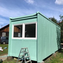GSW Afbouw - Pomalowany na miętowo blaszany pawilon z białymi oknami, ustawiony na pniakach. Aluminiowa drabina oparta o ścianę. W tle dom z ciemnego drewna i szary samochód.