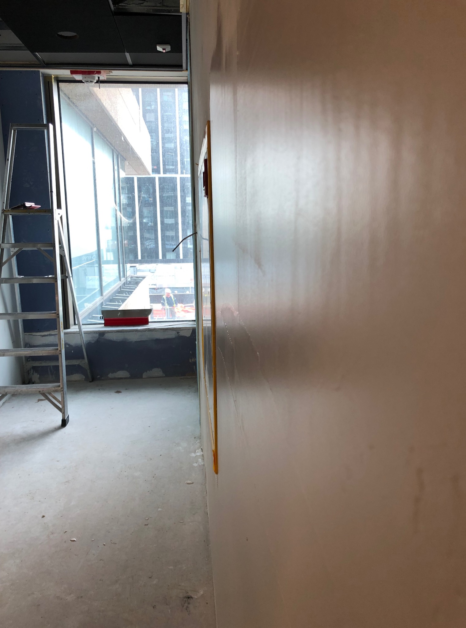 Ściana po szpachlowaniu, widoczne ślady po wałku. W tle okno z widokiem na budynek i drabina. Przygotowanie do malowania w nowoczesnym biurze.