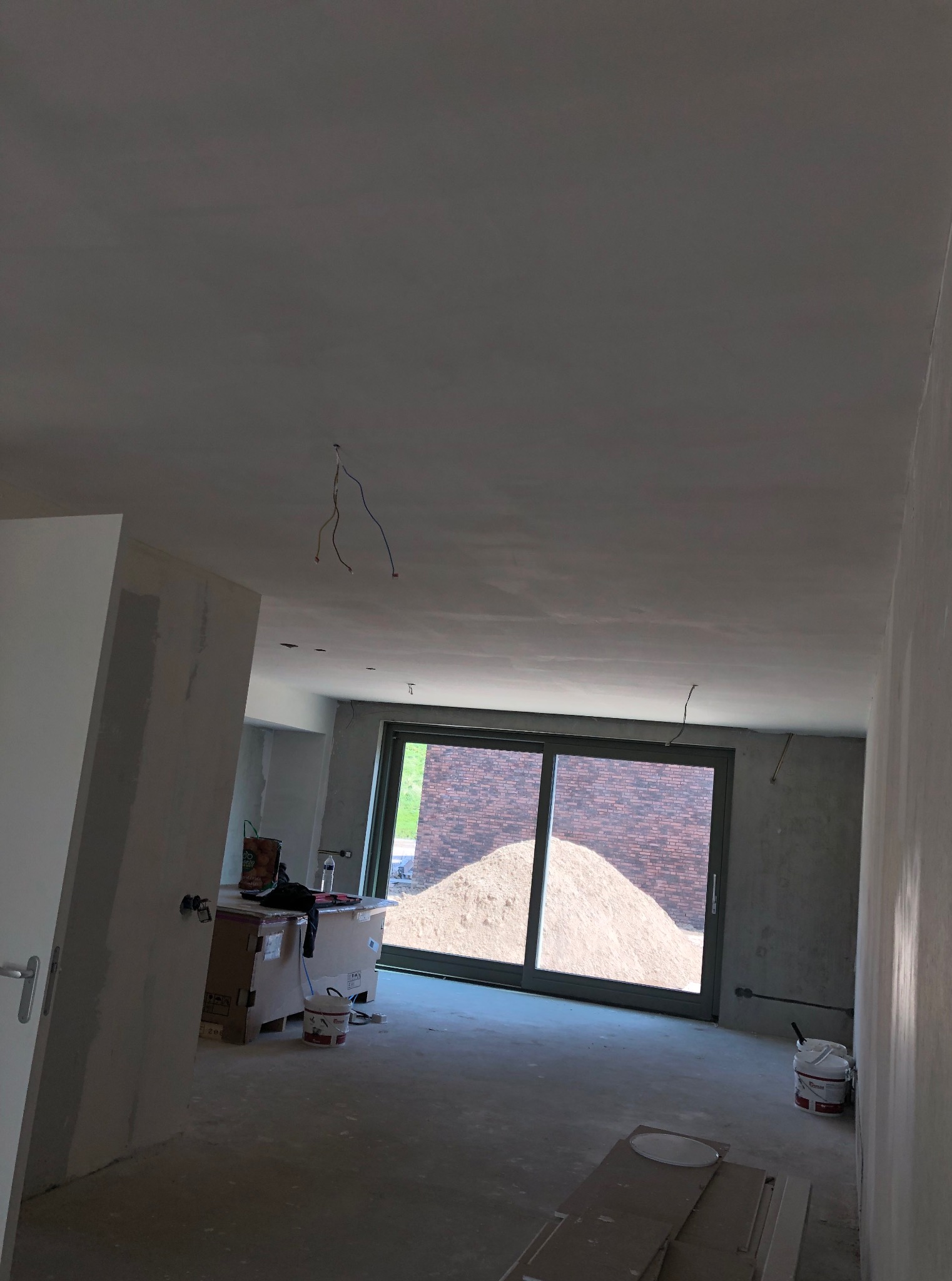 Wnętrze w trakcie prac wykończeniowych: surowe ściany, gładki sufit z wystającymi kablami, okno z widokiem na hałdę piasku. Widoczne narzędzia i materiały.