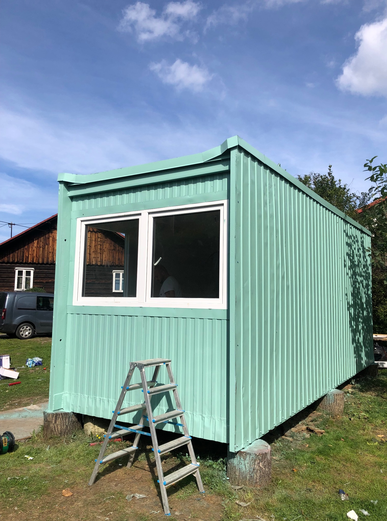 Pomalowany na miętowo blaszany pawilon z białymi oknami, ustawiony na pniakach. Aluminiowa drabina oparta o ścianę. W tle dom z ciemnego drewna i szary samochód.