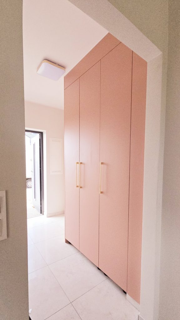 Różowa szafa wnękowa z złotymi uchwytami, w jasnym przedpokoju z białymi płytkami. Minimalistyczny design, widok z perspektywy wejścia do pomieszczenia.
