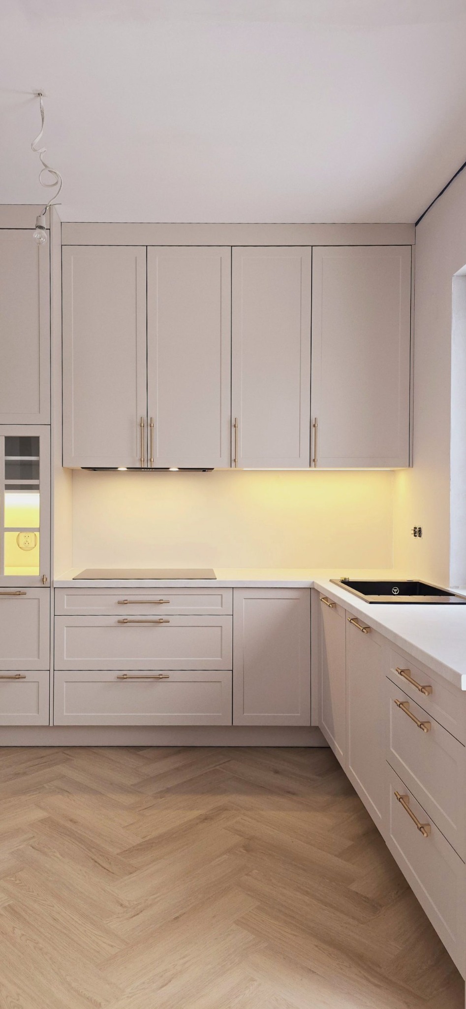 Jasna kuchnia w stylu klasycznym z drewnianą podłogą w jodełkę. Złote uchwyty szafek i szuflad, oświetlenie podszafkowe. Minimalistyczny design.
