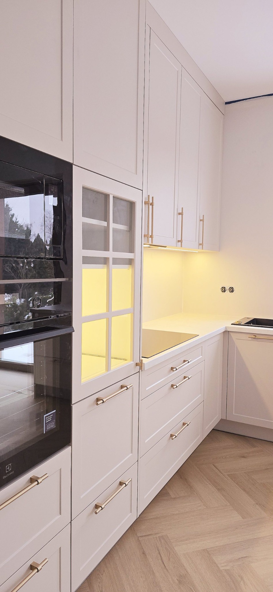 Jasna zabudowa kuchenna z piekarnikiem i mikrofalówką w zabudowie, złote uchwyty, oświetlenie LED pod szafkami, parkiet w jodełkę. Nowoczesny design.