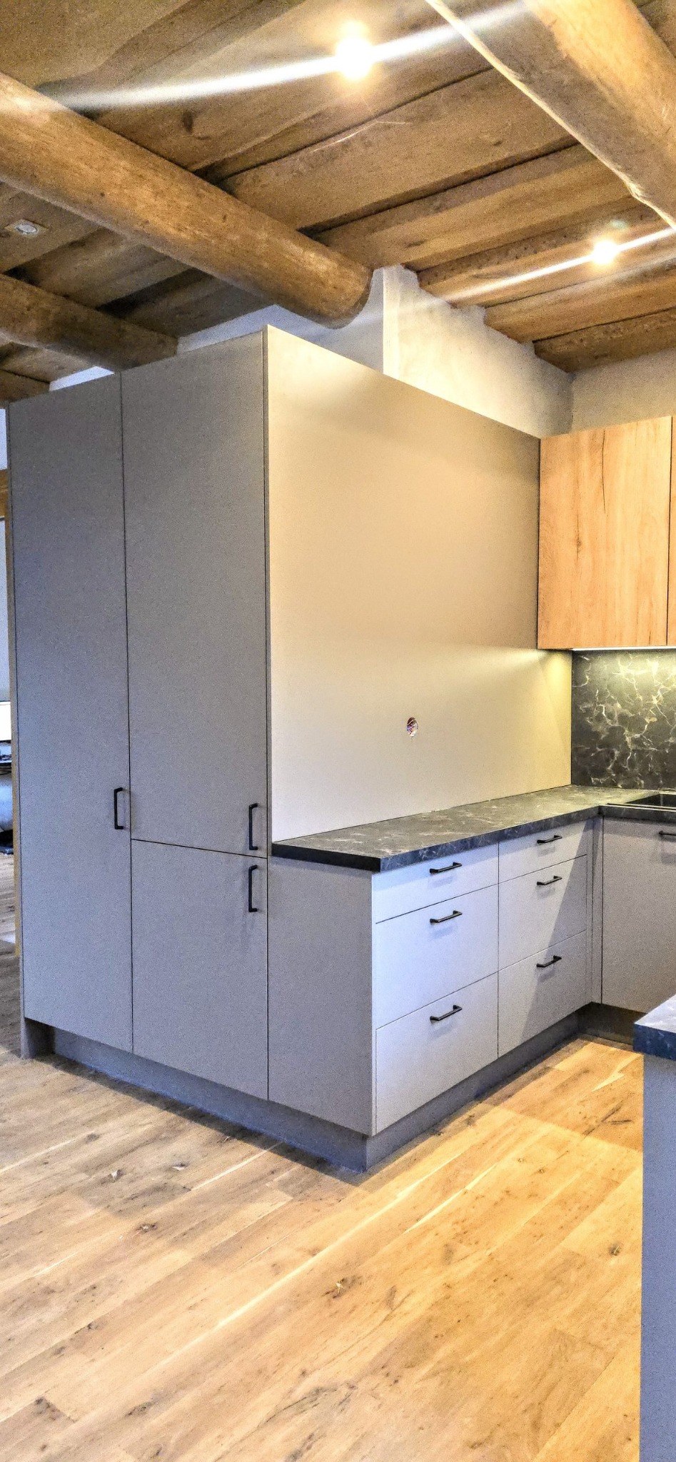 Jasna kuchnia z drewnianym sufitem i czarnymi uchwytami. Szafki dolne z szufladami, górne z drewnianymi frontami. Blat z ciemnego kamienia.