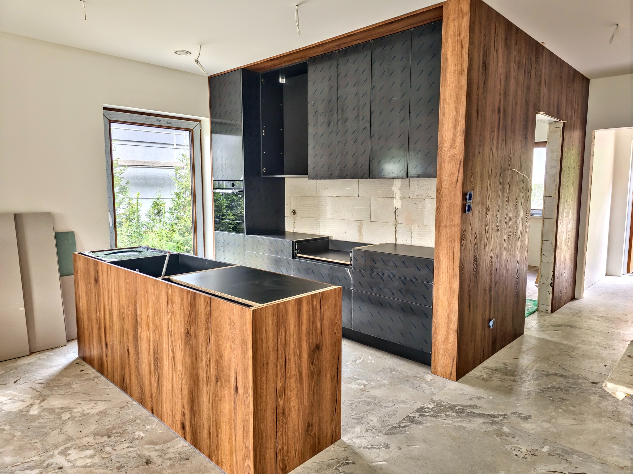 Częściowo zmontowana kuchnia na wymiar z drewnianą wyspą i czarnymi szafkami, widoczne bloki konstrukcyjne i folia ochronna na frontach, surowy stan wykończenia wnętrza.