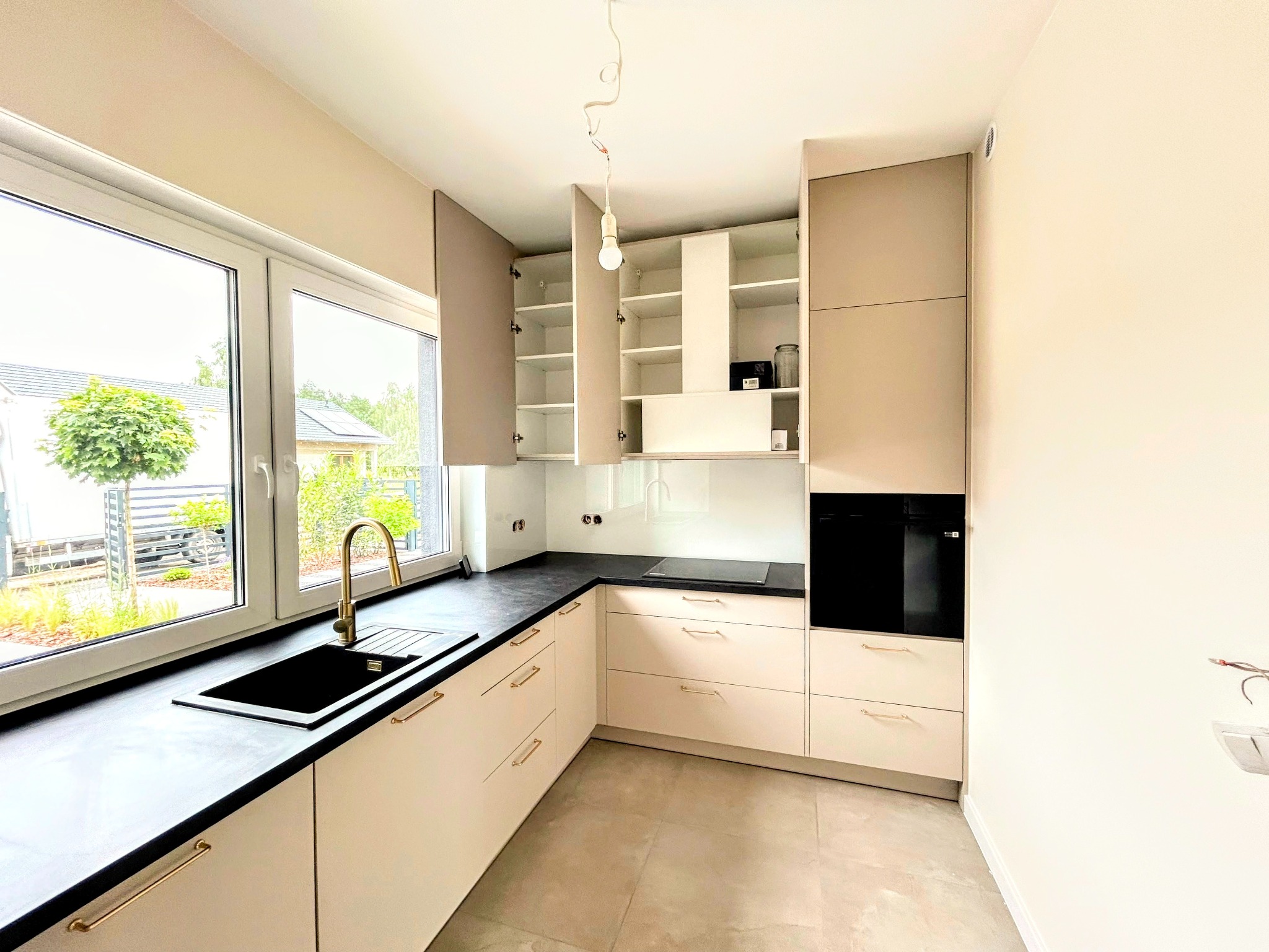 Jasna kuchnia w nowoczesnym stylu z czarnym blatem i złotą armaturą. Widoczne otwarte szafki górne i okno z widokiem na zieleń. Minimalistyczny design.