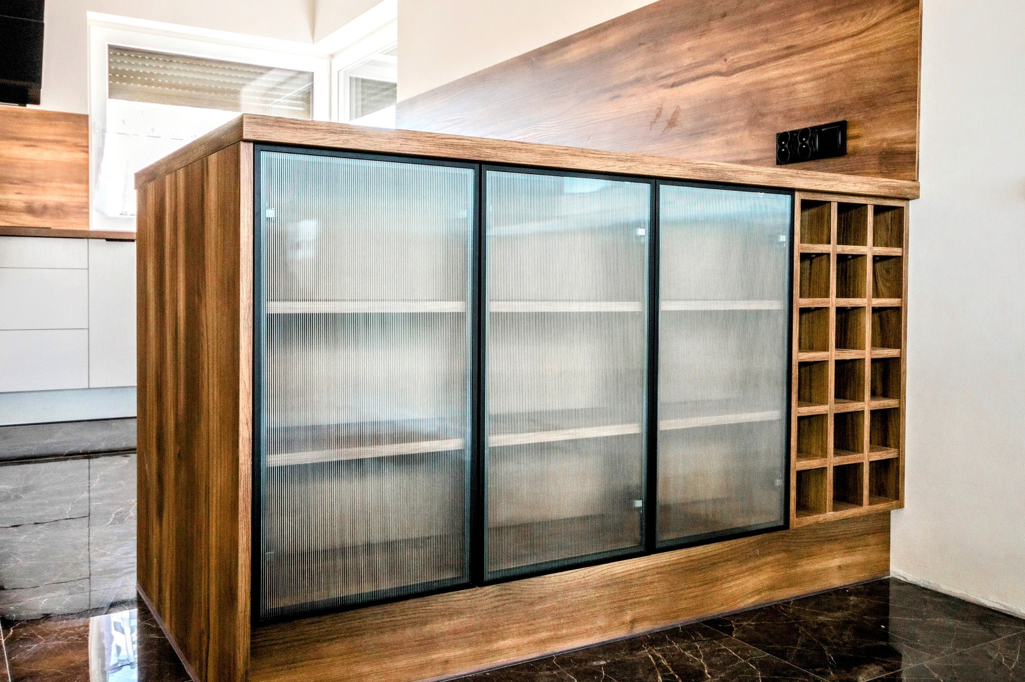 Drewniana szafka z ryflowanymi szklanymi drzwiami i półkami na wino, wkomponowana w drewnianą zabudowę ściany, na tle marmurowej podłogi.