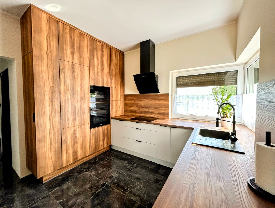 Nowoczesna kuchnia z drewnianymi frontami szafek, czarnym okapem i marmurową podłogą. Widok na okno z roletą i rośliną doniczkową.