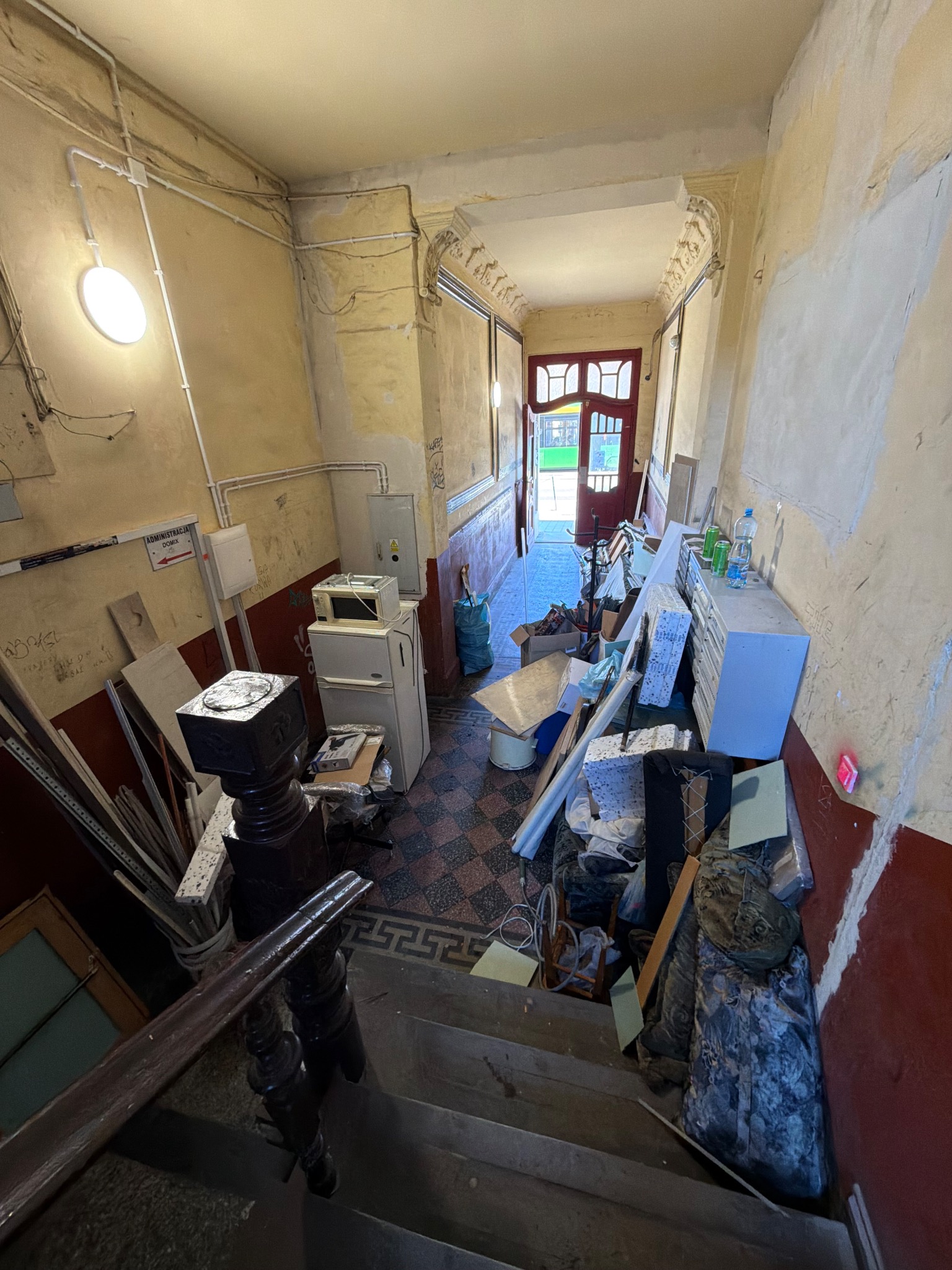 Korytarz w kamienicy w trakcie remontu: widoczne ślady po skuciu tynku, stare instalacje, gruz i sprzęty. Perspektywa z góry schodów.
