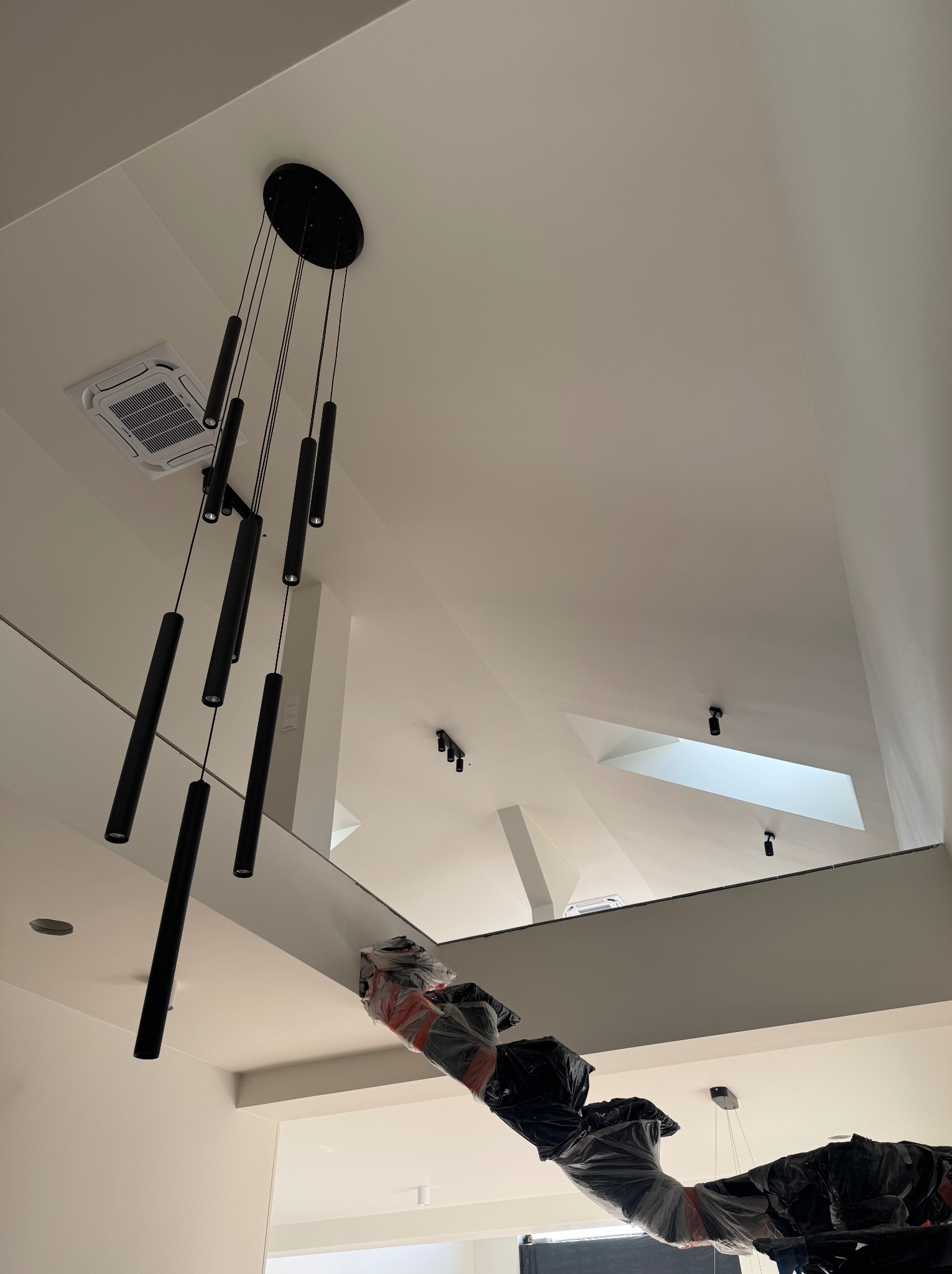 Nowoczesny, czarny żyrandol tubowy zamontowany na białym suficie, widok z dołu. W tle widoczne inne punkty oświetleniowe i klimatyzacja.