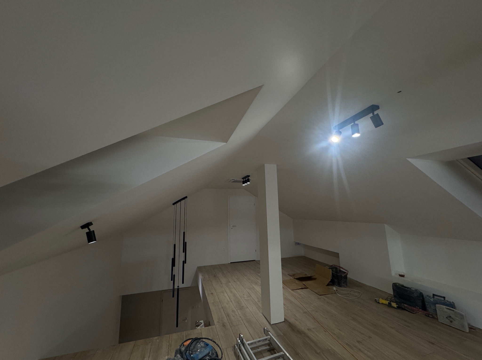 Zamontowane czarne lampy sufitowe w poddaszu ze skosami. Widoczne narzędzia pracy na podłodze. Oświetlenie punktowe i wiszące. Białe ściany i drewniana podłoga.