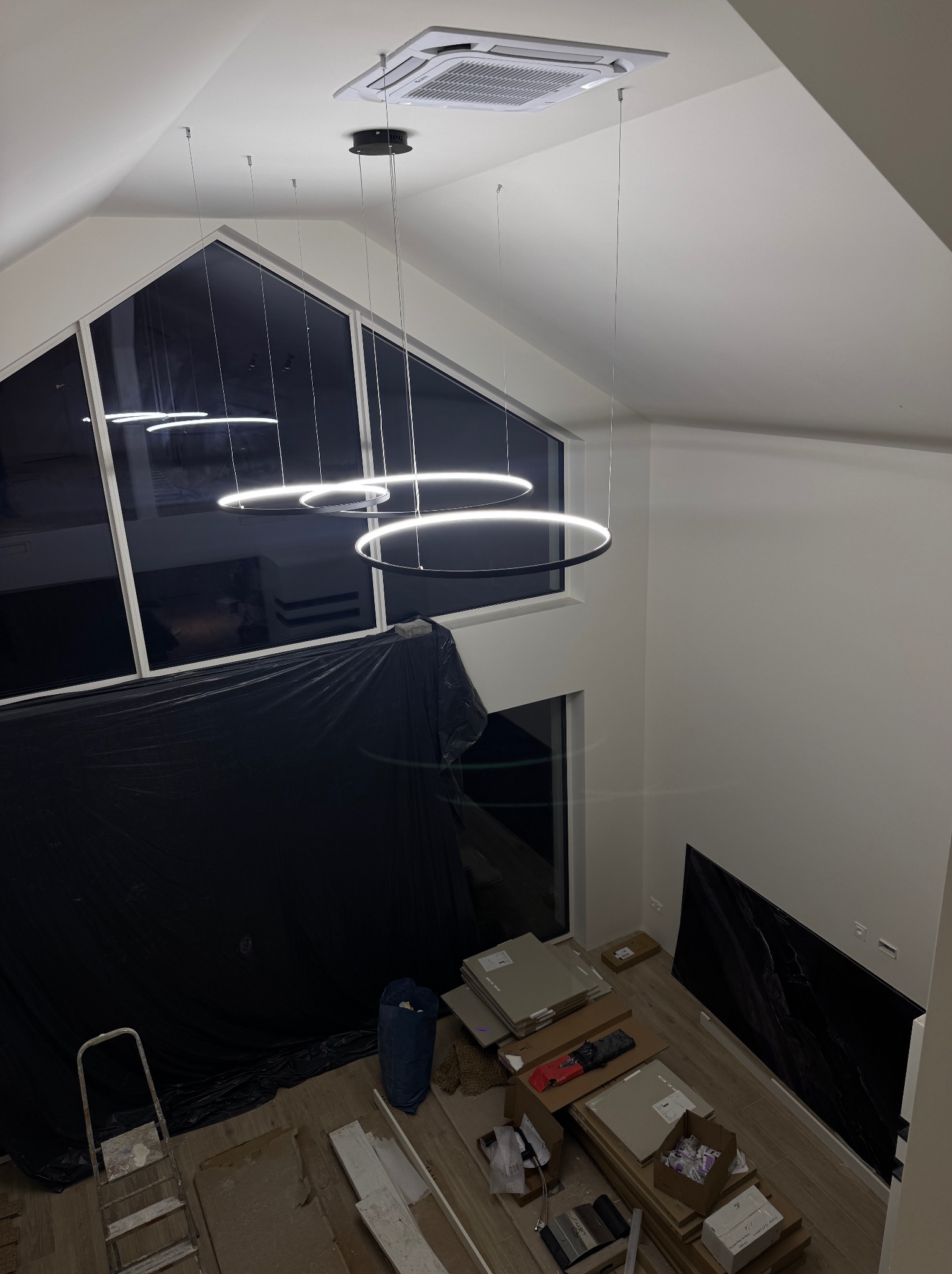 Nowoczesne, okrągłe lampy LED zawieszone w wysokim salonie z dużymi oknami. Widoczne kable montażowe, drabina i materiały budowlane, sugerujące etap instalacji.