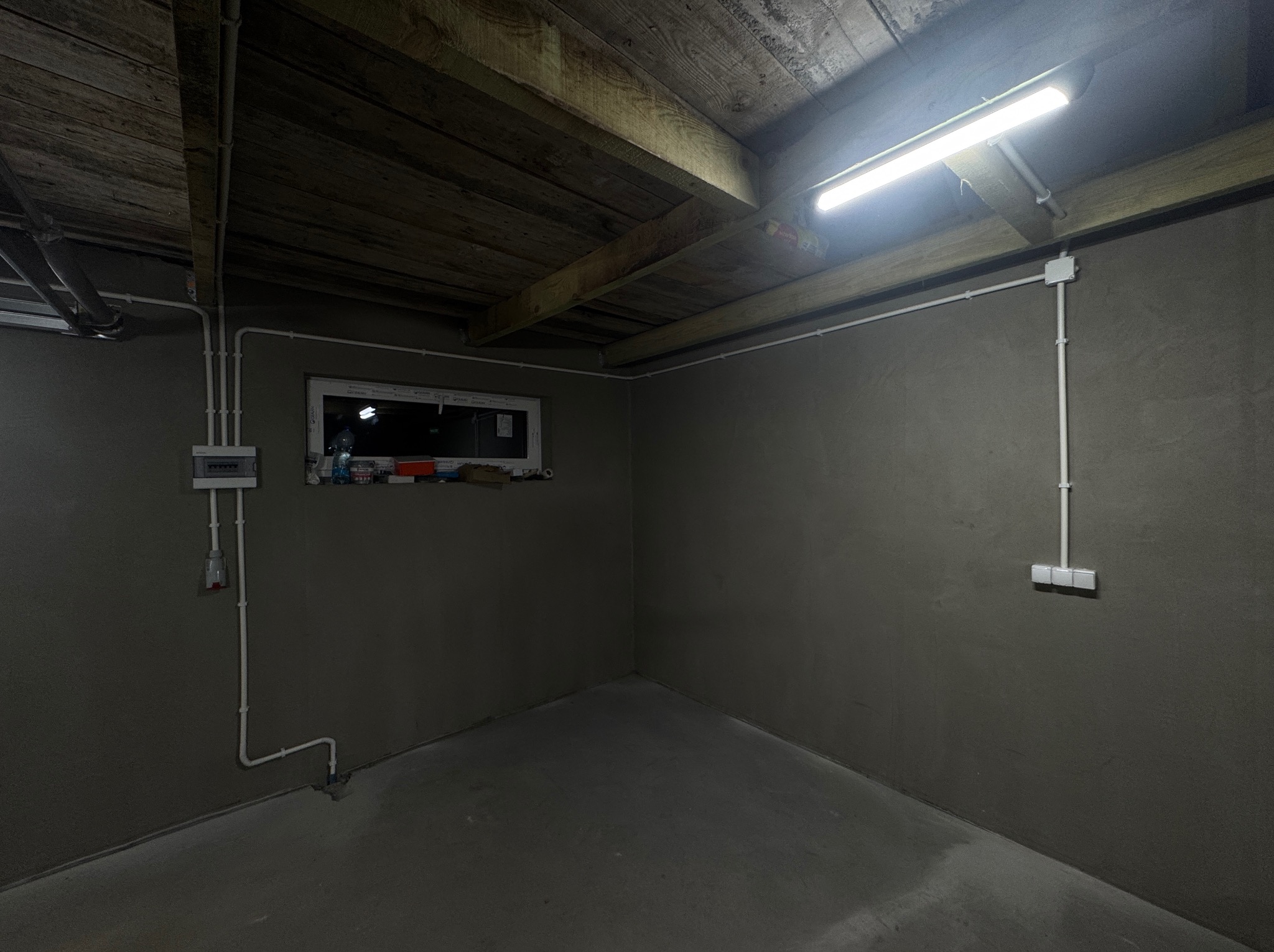 Szara piwnica z oknem i nową instalacją elektryczną: białe kable prowadzone po ścianach, skrzynka rozdzielcza, gniazdka i lampa LED pod drewnianym stropem.