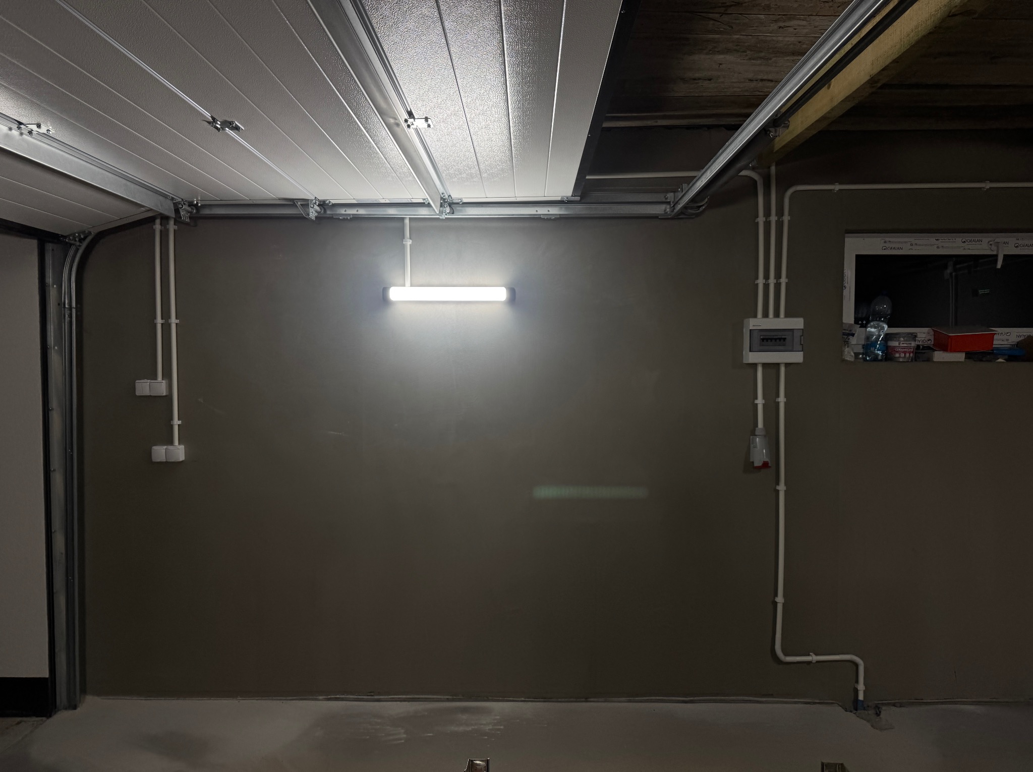 Zamontowana lampa LED na ścianie garażu z widoczną instalacją elektryczną w białych rurkach. Szara ściana, betonowa podłoga i fragment bramy garażowej.