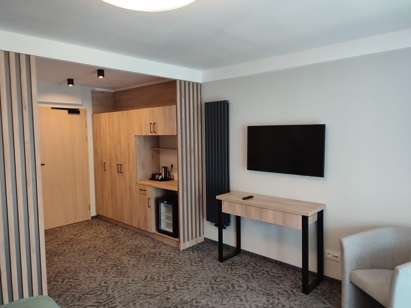 Nowoczesny pokój hotelowy z drewnianą zabudową, biurkiem i telewizorem. Minimalistyczny design, szara kolorystyka, oświetlenie punktowe. Widok na wnętrze pokoju.
