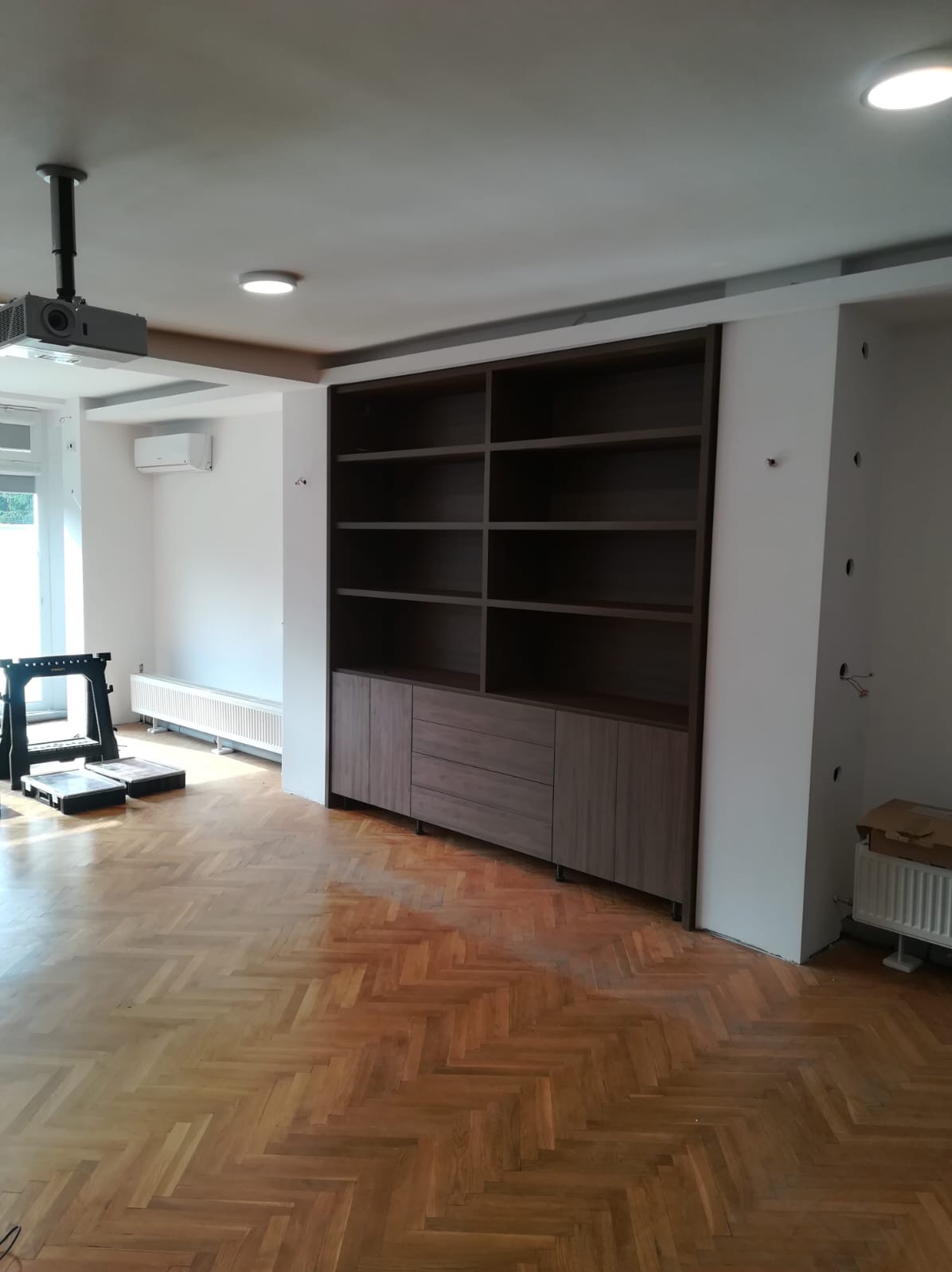 Wnętrze z drewnianą podłogą w jodełkę i zabudowaną szafą z półkami. Widoczny projektor pod sufitem i grzejnik. Ściany białe, z widocznymi otworami na kable.
