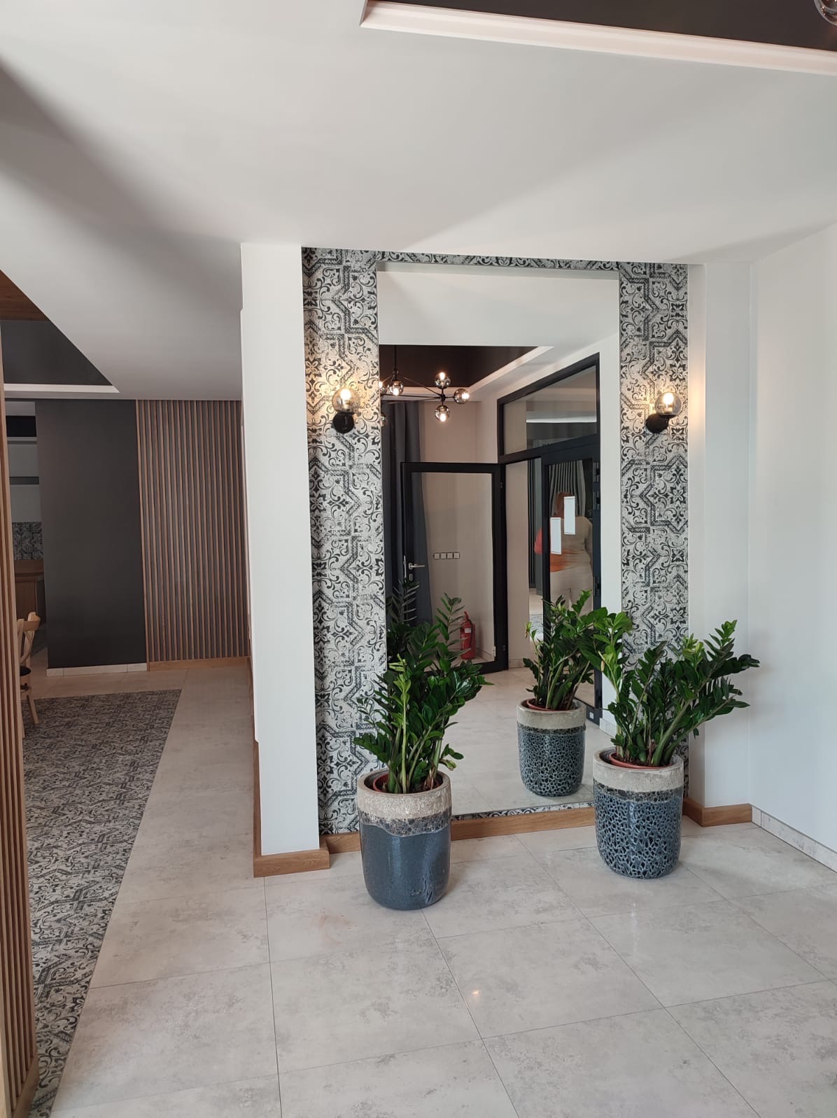 Nowoczesne wnętrze: lustro w dekoracyjnej ramie z wzorem, oświetlone kinkietami, z roślinami w donicach. Widok na korytarz z elementami drewnianymi i dywanem.