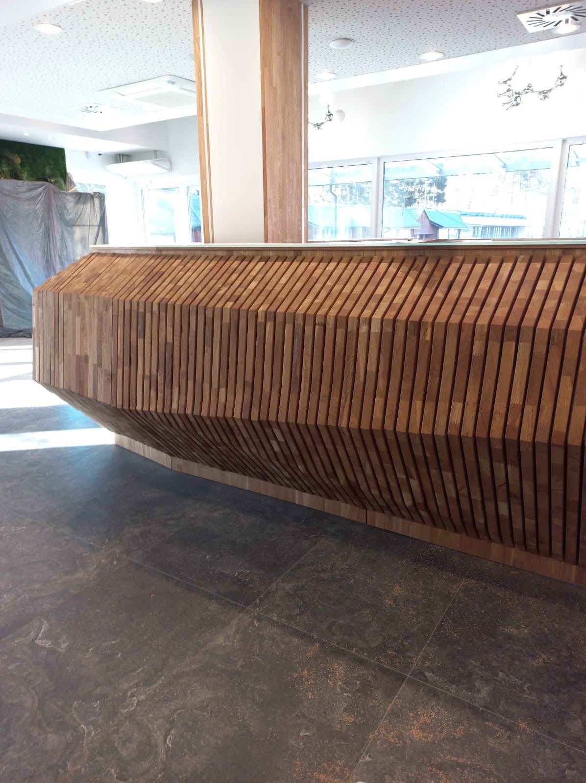 Nowoczesna lada recepcyjna z drewnianych listew, oświetlona naturalnym światłem z okien. Widoczna struktura drewna i ciemna posadzka. Element wykończenia wnętrza.