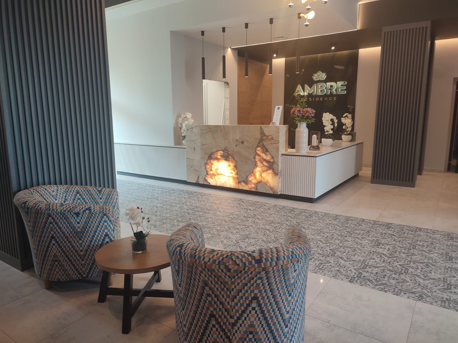 Eleganckie wnętrze recepcji z podświetlaną ladą z kamienia, wzorzystą podłogą i fotelami w geometryczne wzory. Nowoczesne oświetlenie i dekoracje kwiatowe.