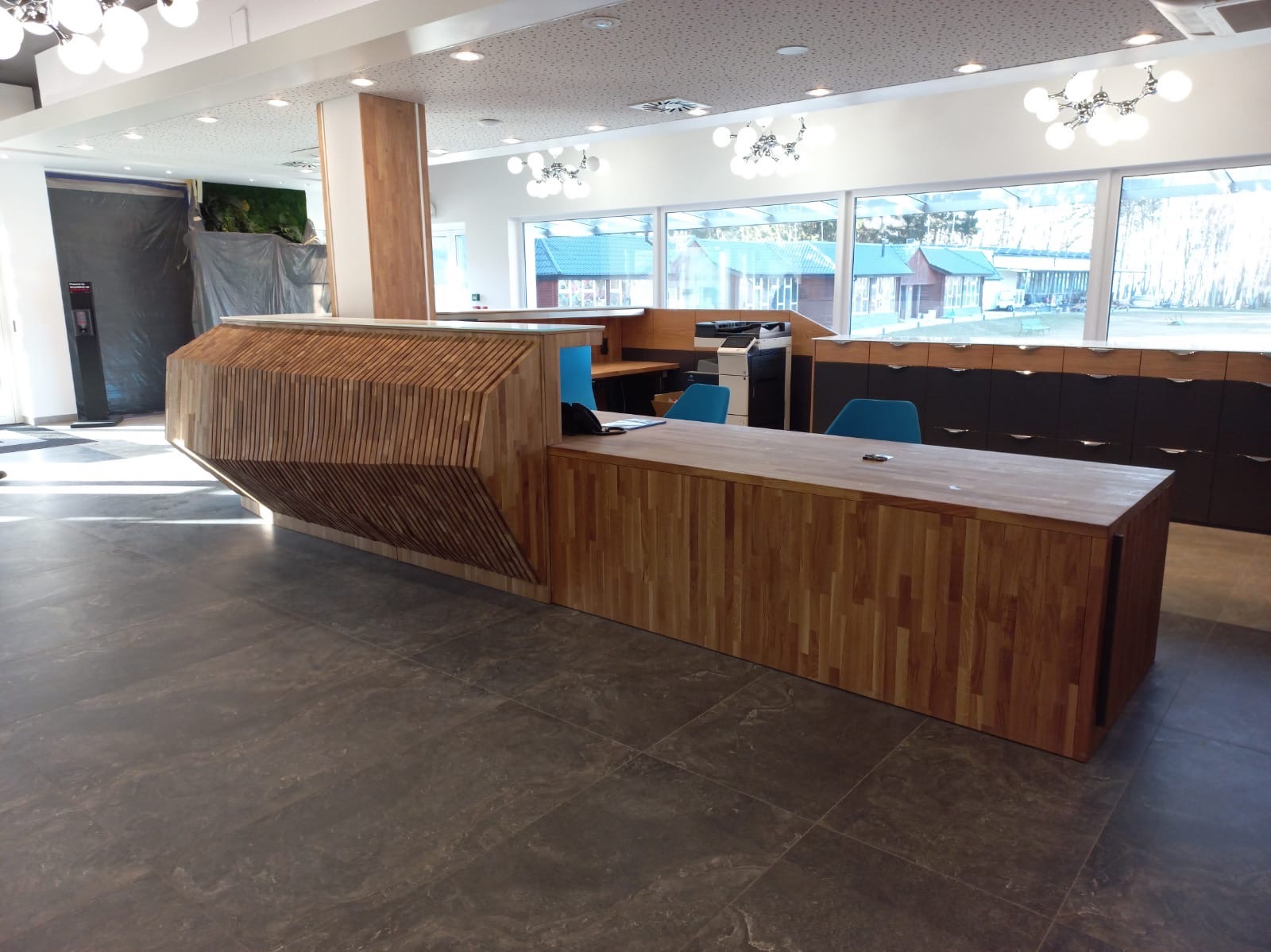 Nowoczesna, drewniana lada recepcyjna z unikalnym designem w przestronnym biurze. Widoczne elementy wykończenia wnętrz i oświetlenie.