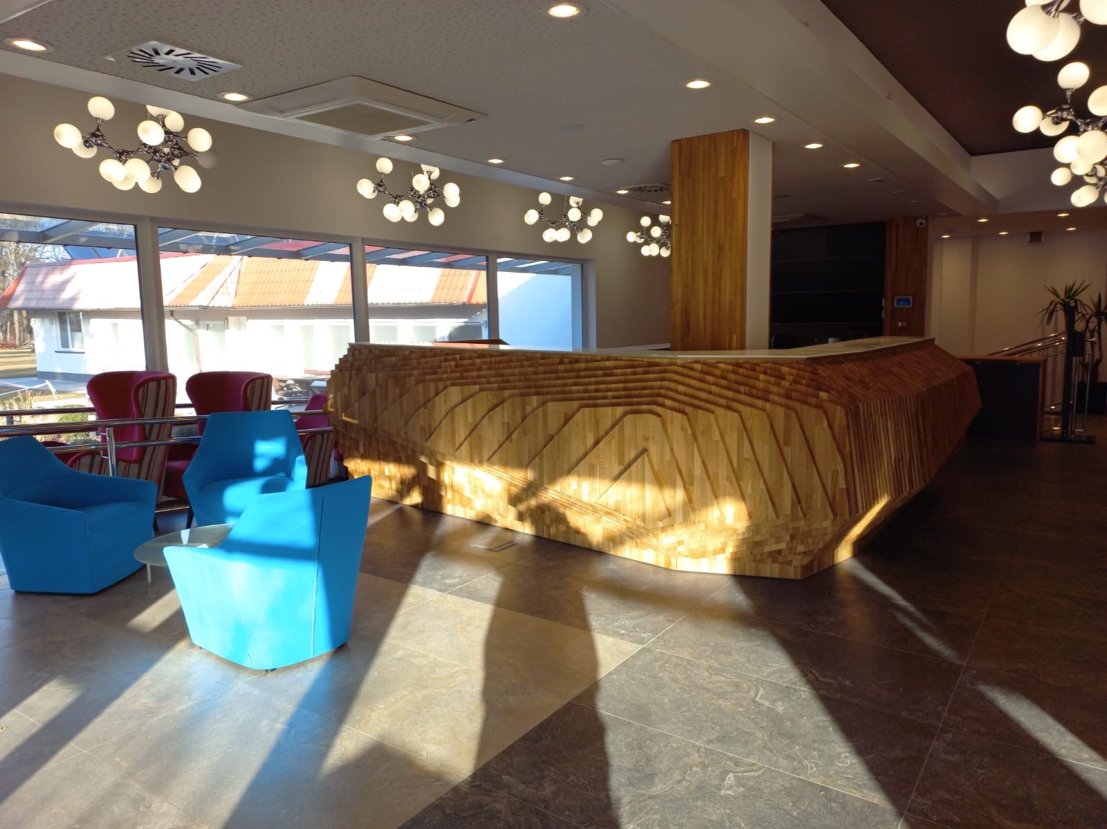 Nowoczesne wnętrze biura z drewnianą recepcją o unikalnym designie, błękitnymi fotelami i dużymi oknami z widokiem na zewnątrz. Oświetlenie punktowe i designerskie lampy.