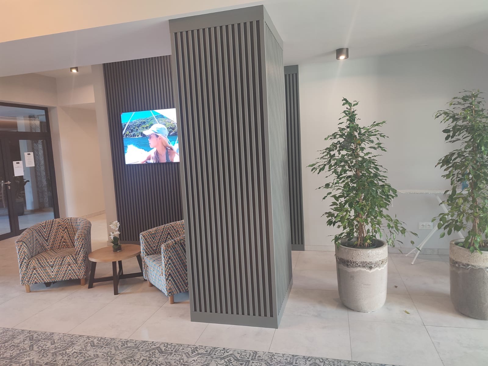 Nowoczesne wnętrze: szare lamele ścienne, TV, wzorzyste fotele, stolik z kwiatami i doniczkowe rośliny. Jasna podłoga z subtelnym wzorem.