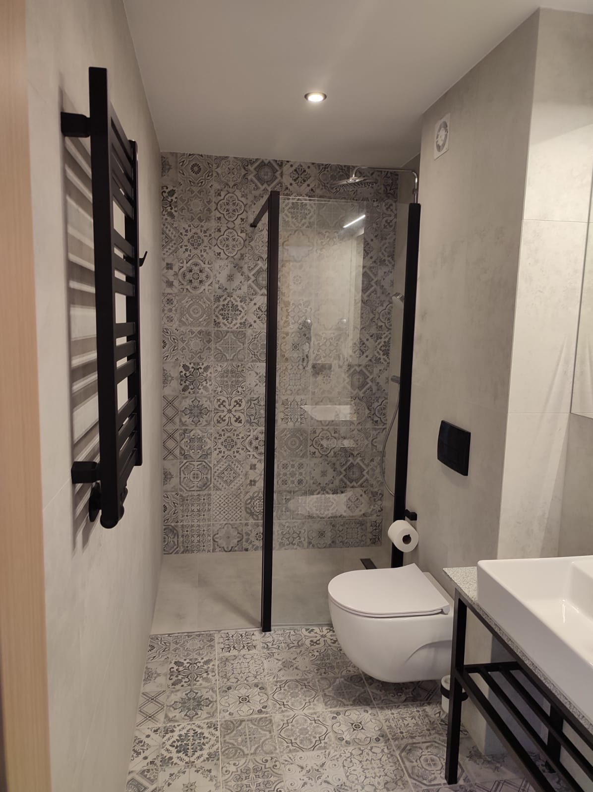 Wykończona łazienka z kabiną prysznicową typu walk-in, wzorzystymi płytkami podłogowymi i ściennymi, oraz czarnym grzejnikiem drabinkowym i armaturą.