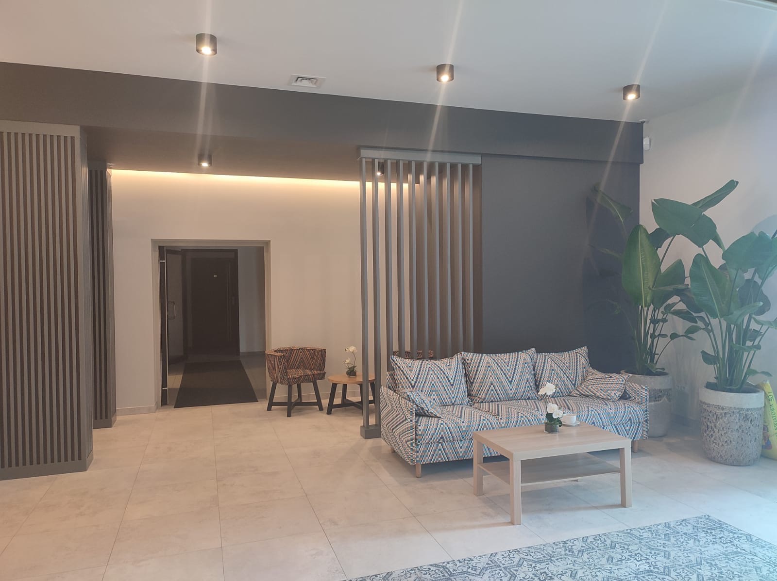 Nowoczesne wykończenie wnętrza: jasna podłoga, sofa w geometryczne wzory, drewniane lamele na ścianie i rośliny doniczkowe. Minimalistyczny design.