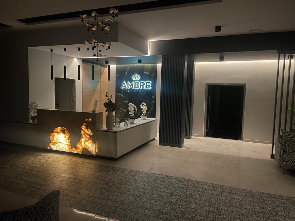 Nowoczesne lobby z podświetlaną recepcją z onyksu i neonowym logo 'Ambre Residence'. Minimalistyczne lampy wiszące i eleganckie wykończenie wnętrz.