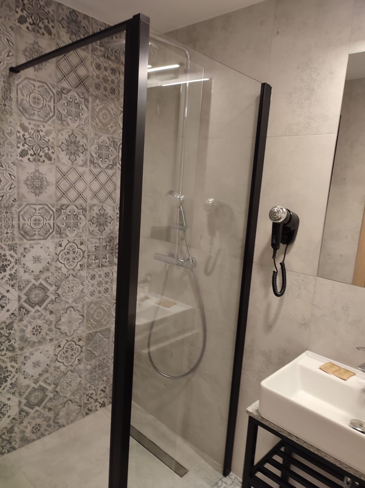 Nowoczesna kabina prysznicowa z czarną ramą i wzorzystymi płytkami w odcieniach szarości. Umywalka z szafką i suszarka do włosów na ścianie.