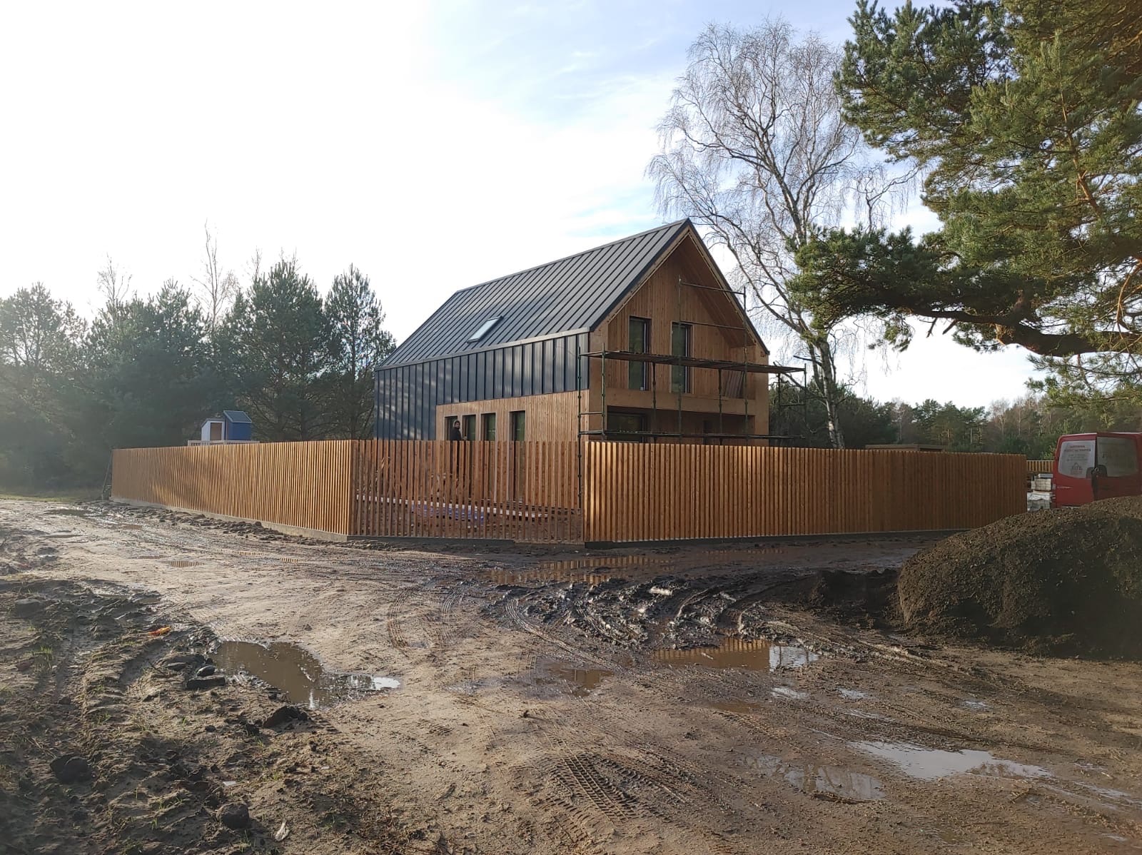 Nowoczesny dom z drewnianą elewacją i metalowym dachem, otoczony nowym, drewnianym ogrodzeniem lamelowym. Teren wokół domu w trakcie prac ziemnych.