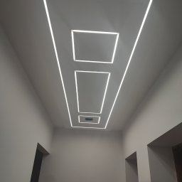 Rs-bud Usługi Remontowo- Wykończeniowe Roman Sądkiewicz - Nowoczesny sufit z oświetleniem LED w kształcie kwadratów i prostokątów, perspektywa od dołu, widoczne fragmenty ścian i drzwi.