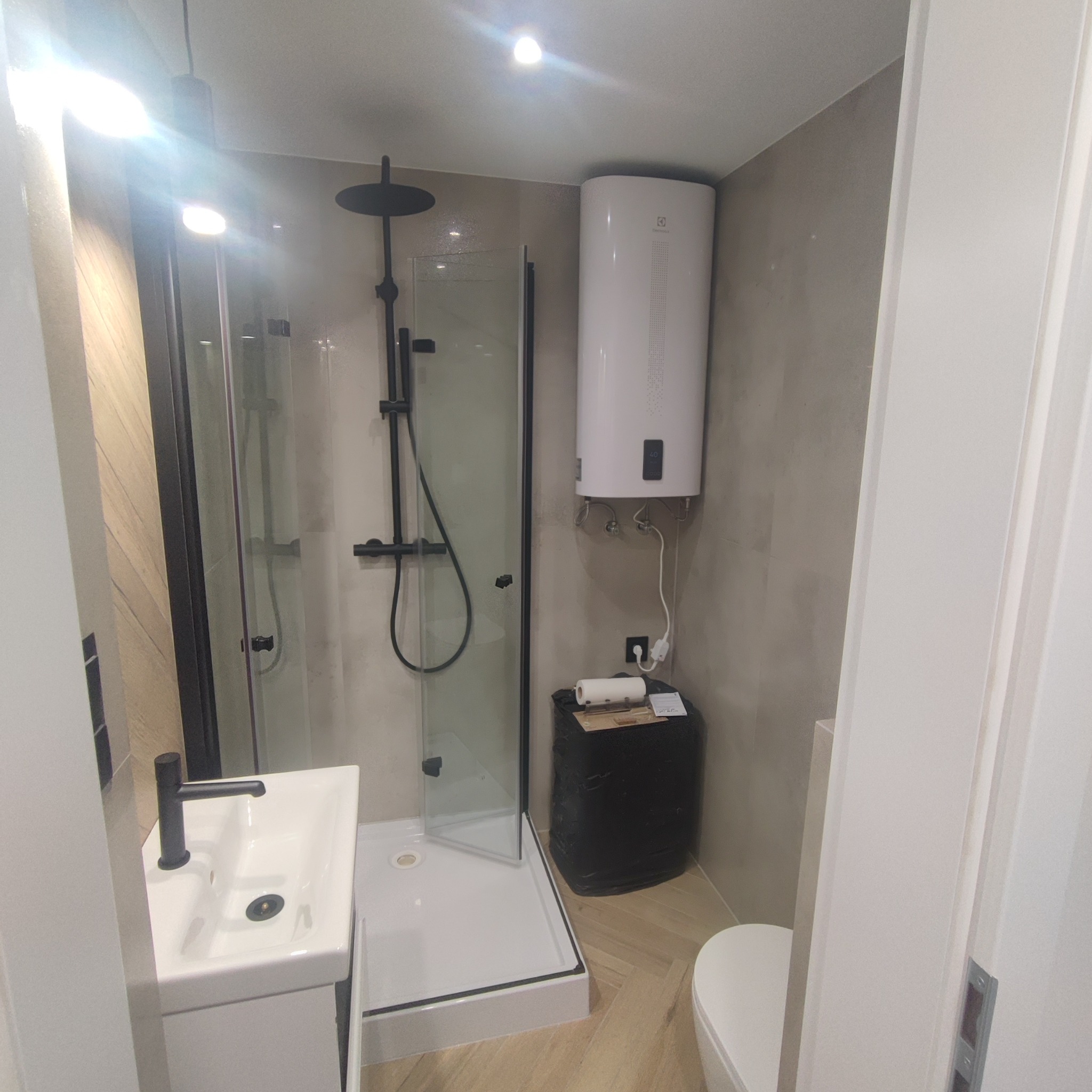 Nowoczesna łazienka po remoncie: kabina prysznicowa typu walk-in z czarną armaturą, biała umywalka i bojler. Ściany wykończone w odcieniach szarości, podłoga imitująca drewno.