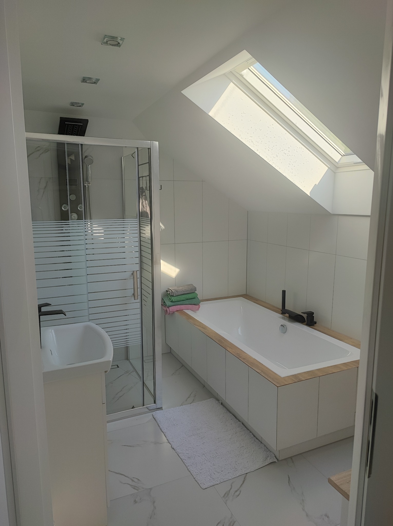 Jasna łazienka na poddaszu z kabiną prysznicową, wanną obudowaną drewnem i białymi płytkami. Okno dachowe wpuszcza naturalne światło. Minimalistyczny design.