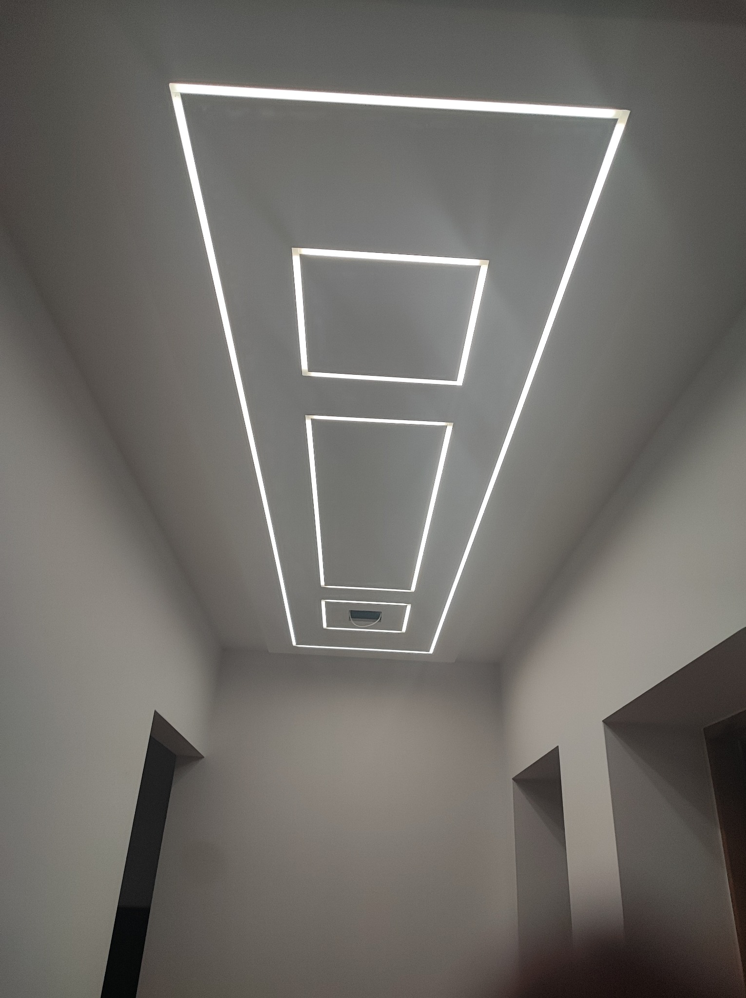 Nowoczesny sufit z oświetleniem LED w kształcie kwadratów i prostokątów, perspektywa od dołu, widoczne fragmenty ścian i drzwi.