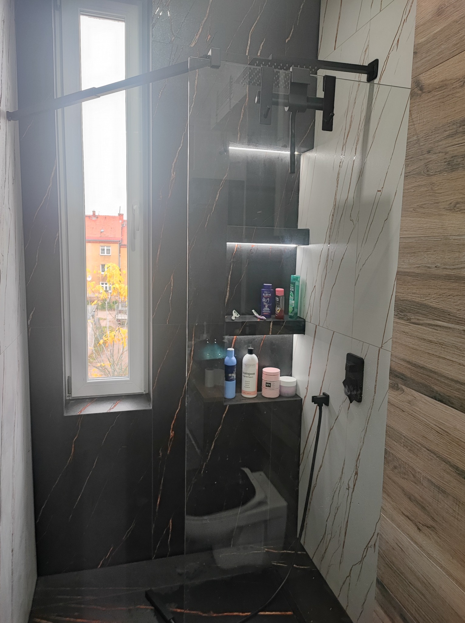 Nowoczesna kabina prysznicowa z czarnymi elementami i marmurowymi płytkami, widok na okno z jesiennym krajobrazem. Minimalistyczny design, eleganckie wykończenie.