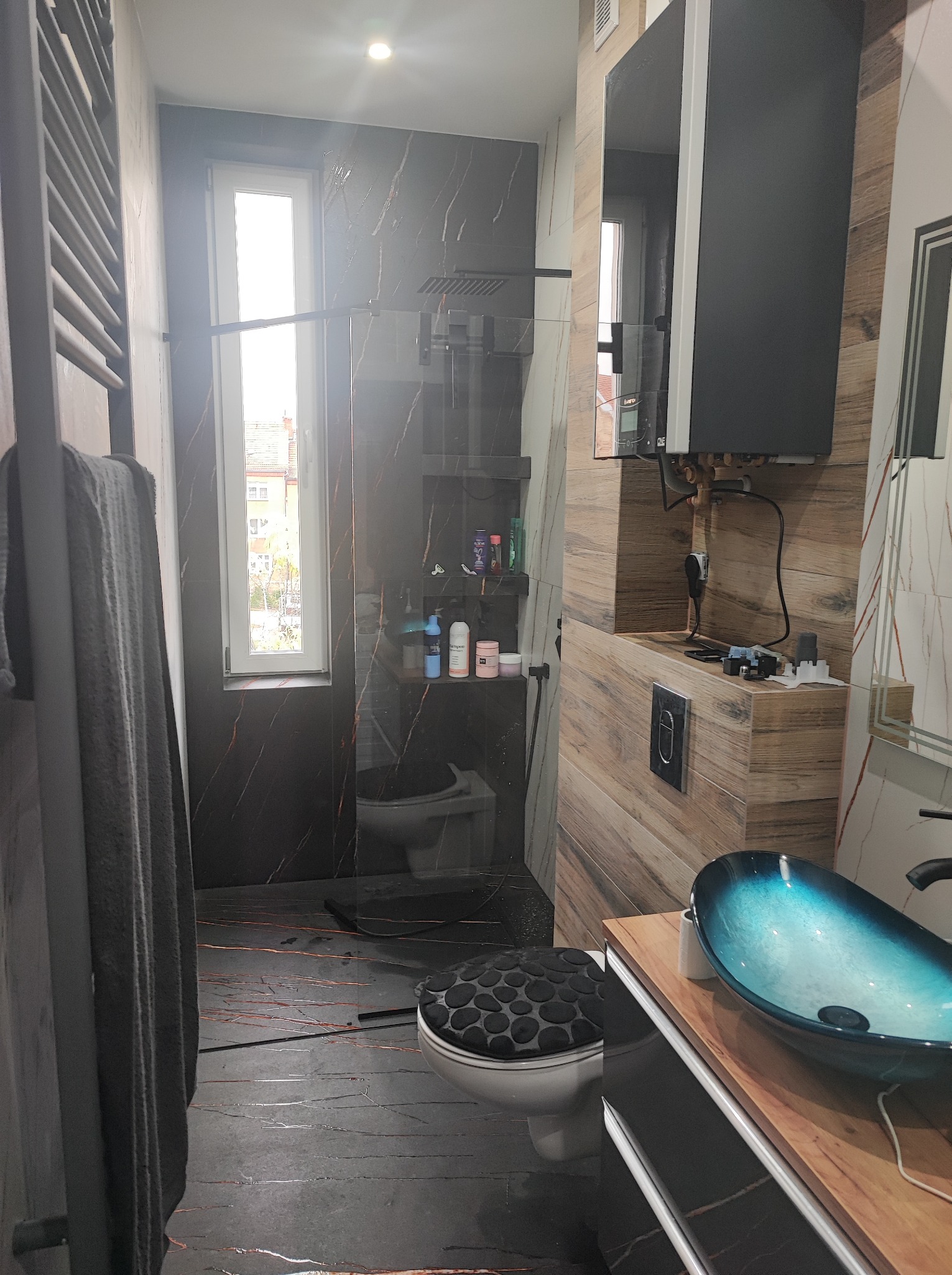 Nowoczesna łazienka z czarnymi płytkami imitującymi marmur, szklaną kabiną prysznicową i umywalką w odcieniach błękitu. Widoczny grzejnik drabinkowy i drewnopodobne wykończenia.