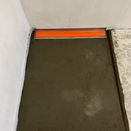 Mateusz Pakuła - Wyrównywanie wylewki podłogowej w rogu pomieszczenia za pomocą pomarańczowej łaty. Widoczny fragment ściany i surowa podłoga.