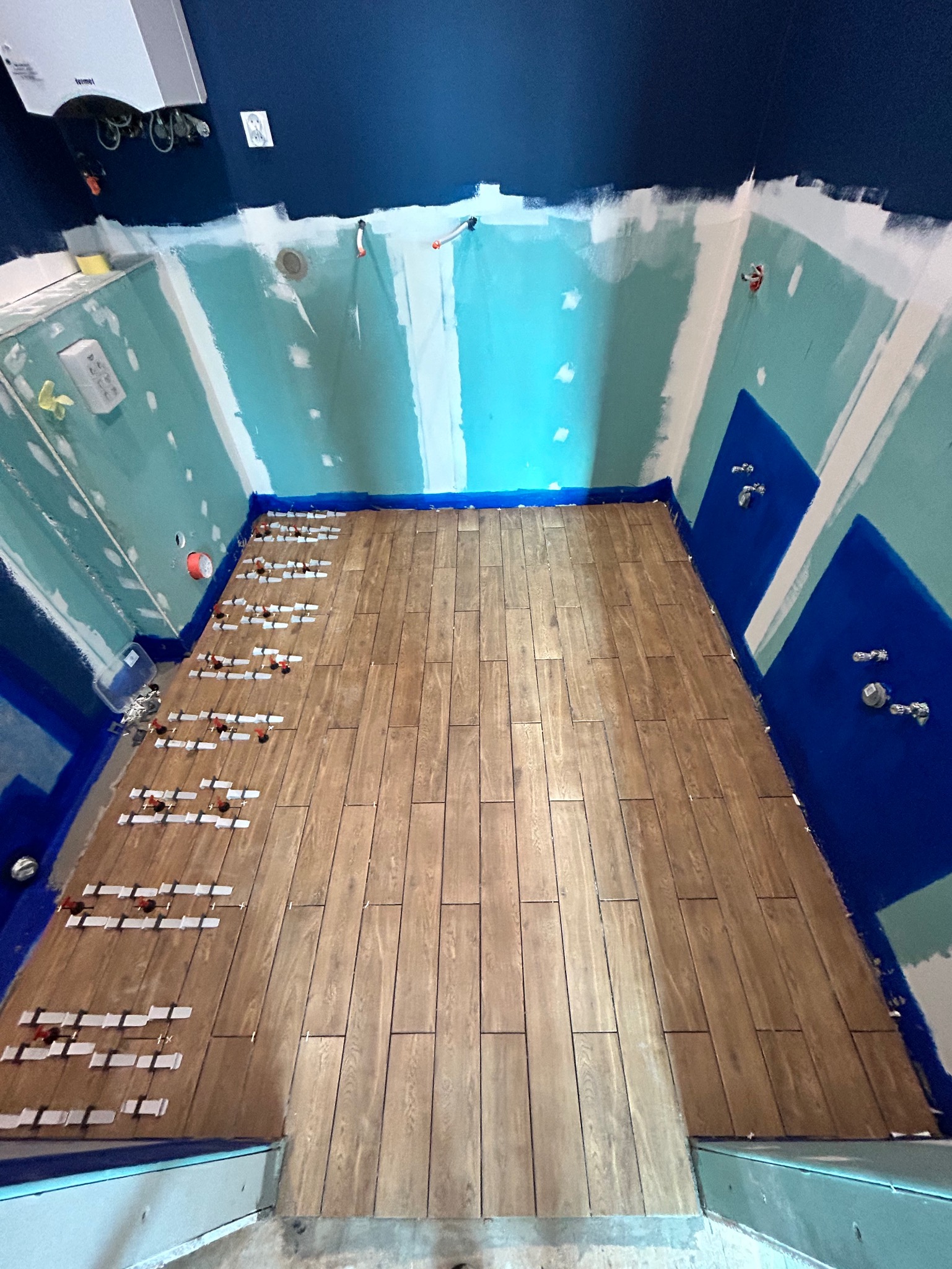 Układanie drewnopodobnych płytek podłogowych w łazience z przygotowanymi ścianami pod glazurę. Widoczne narzędzia do poziomowania płytek.