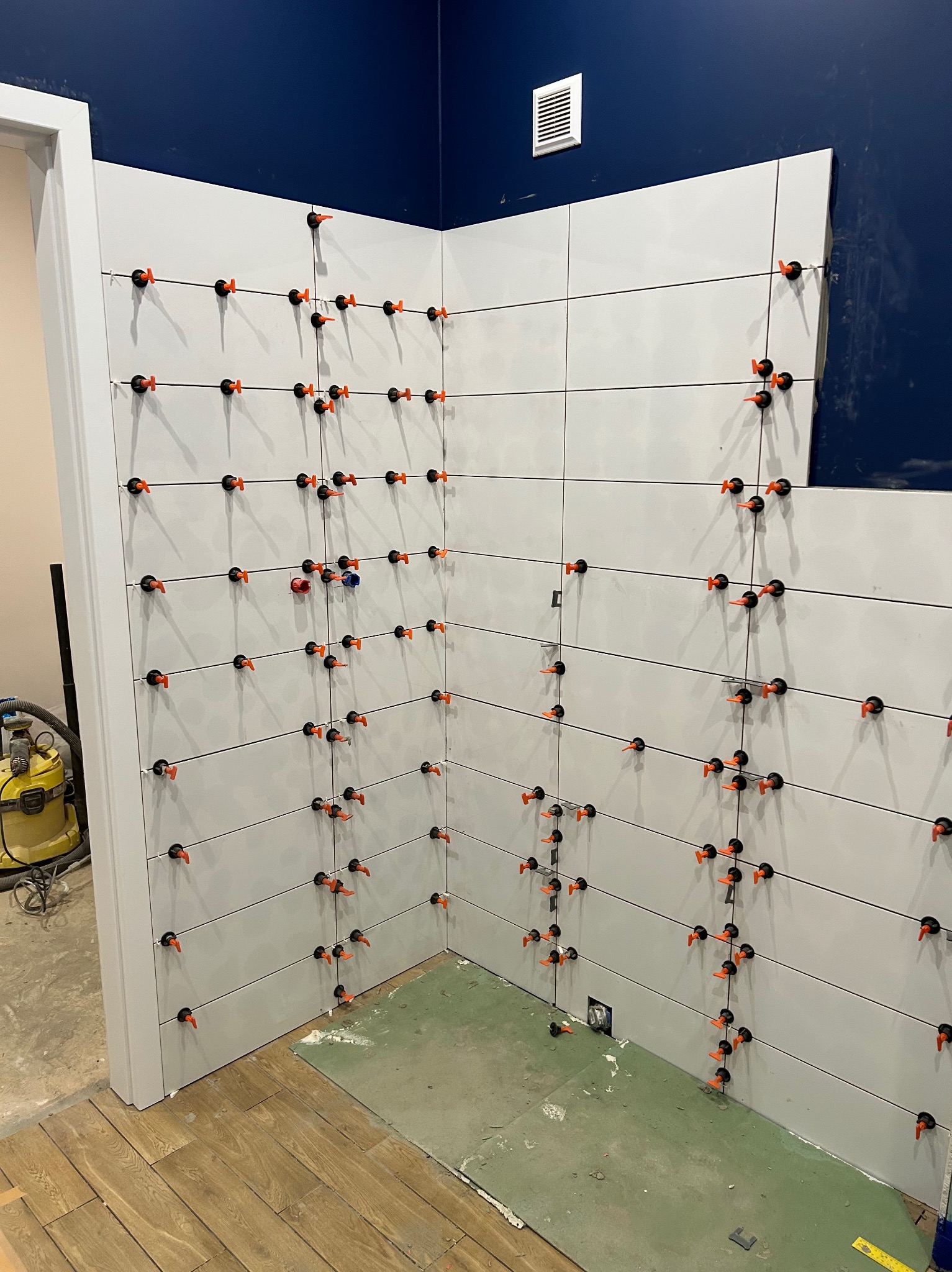 Świeżo ułożone, białe płytki na ścianie z systemem poziomowania, widoczne narzędzia i fragment podłogi. Etap prac wykończeniowych w łazience.