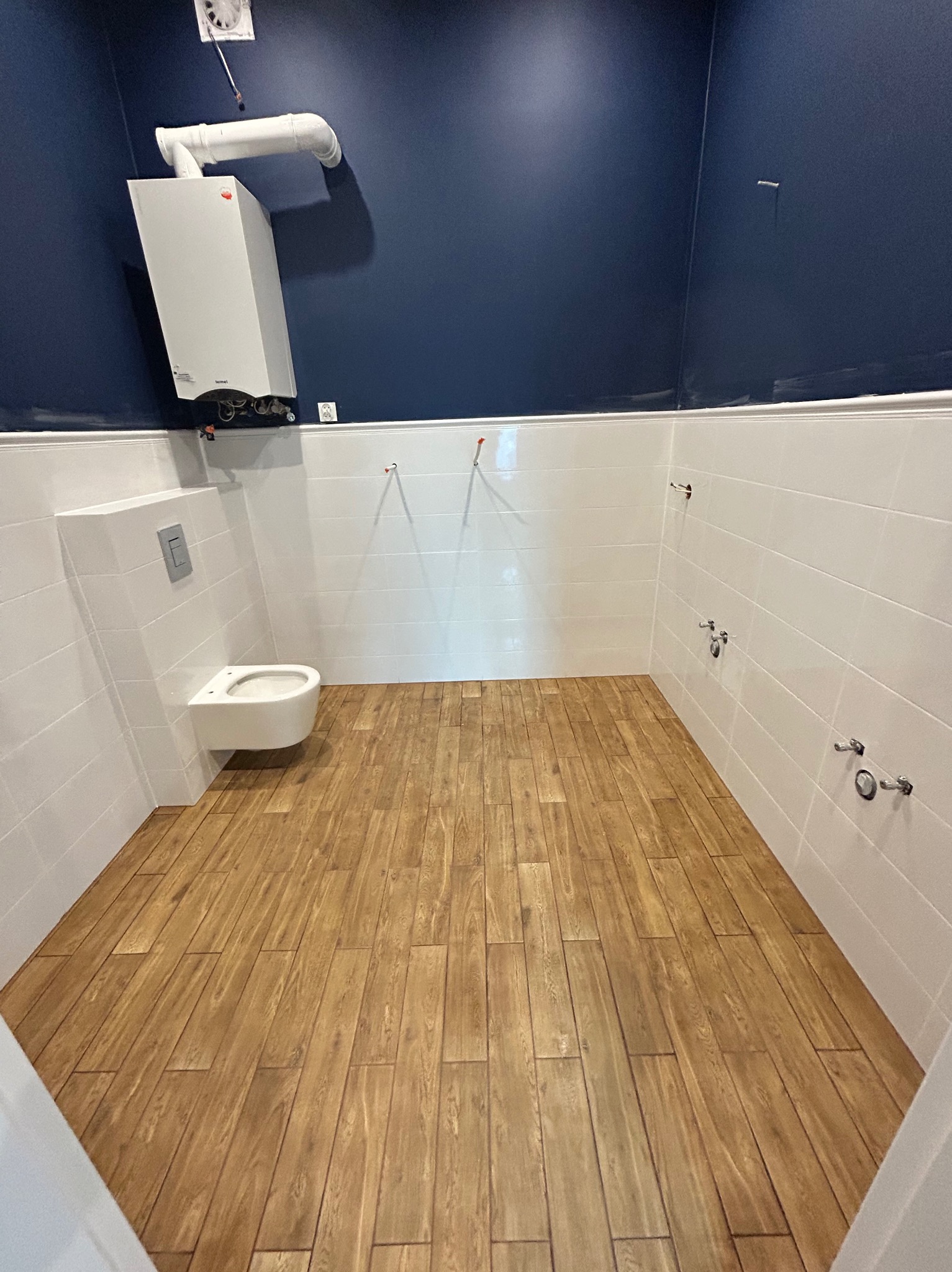 Wykończona łazienka z drewnopodobną podłogą, białymi płytkami na ścianach i wiszącą toaletą. Na ścianie ciemnoniebieska farba i piec gazowy.