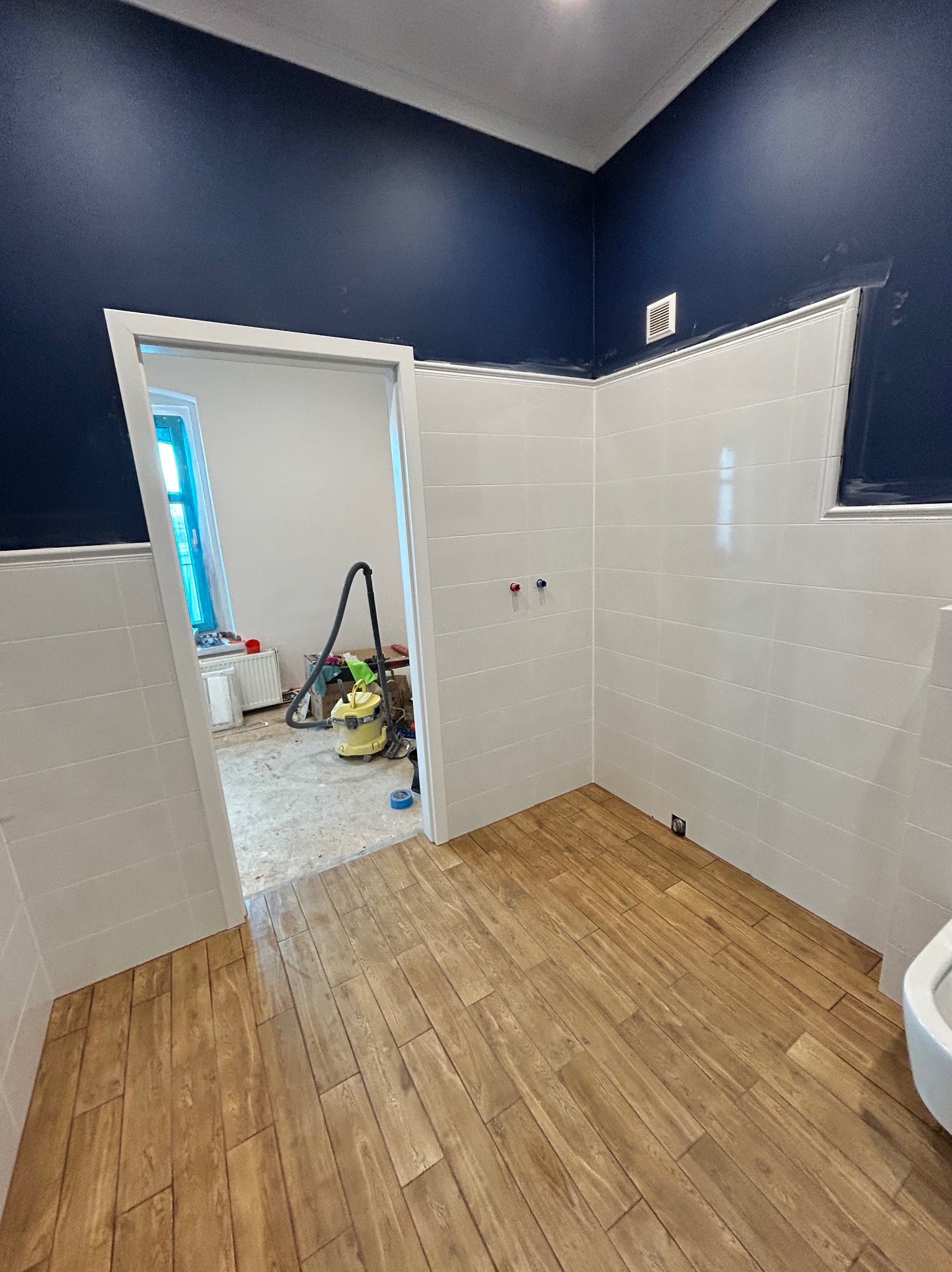 Wykończona łazienka z drewnopodobną podłogą i białymi płytkami na ścianach. Widoczne niebieskie ściany i odkurzacz w sąsiednim pomieszczeniu.